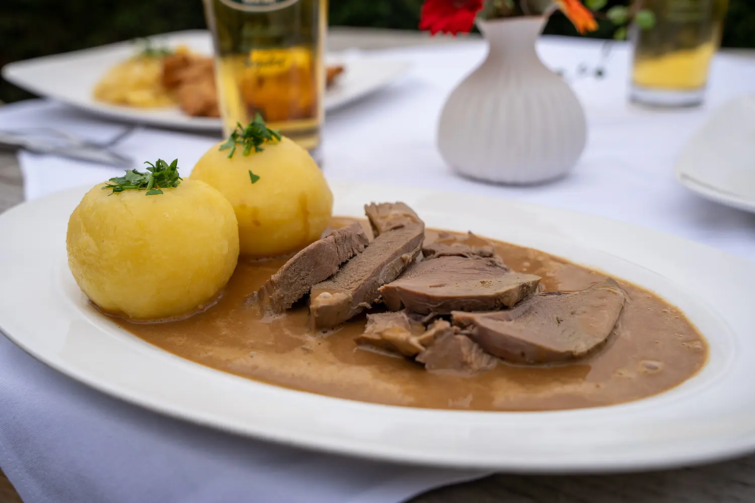 Rehbraten in Scheiben mit brauner Sauce und zwei mit Kräutern bestreuten Kartoffelklößen.