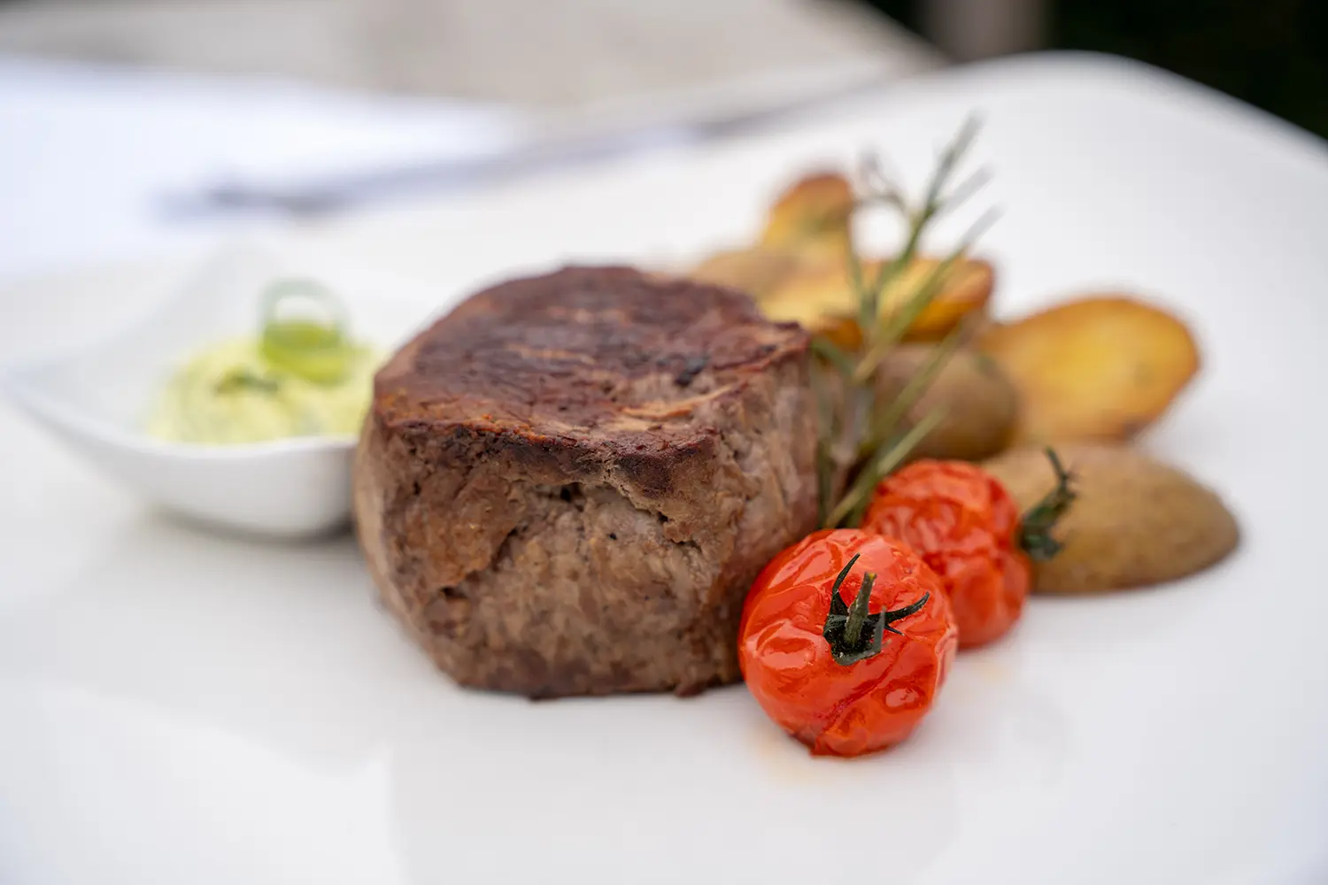 Gebratenes Steak mit Rosmarinkartoffeln, kleinen Tomaten und einem Schälchen Kräuterbutter.